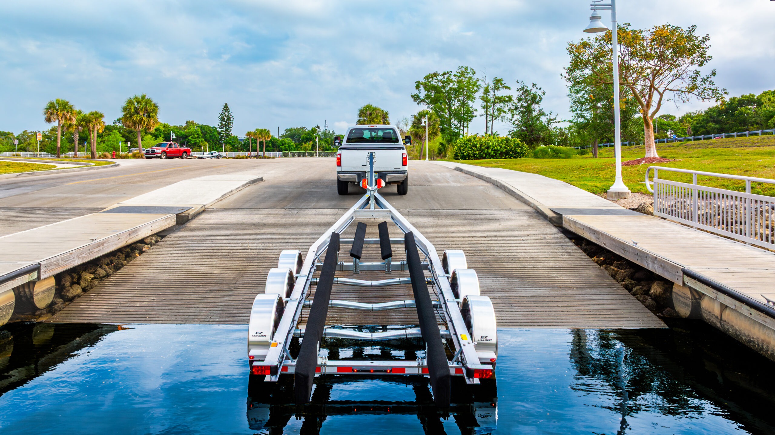 EZ Loader aluminum boat trailer backed into boat ramp by white pickup truck, showing quad bunk system, carpeted supports, and reflective safety lighting at launch site