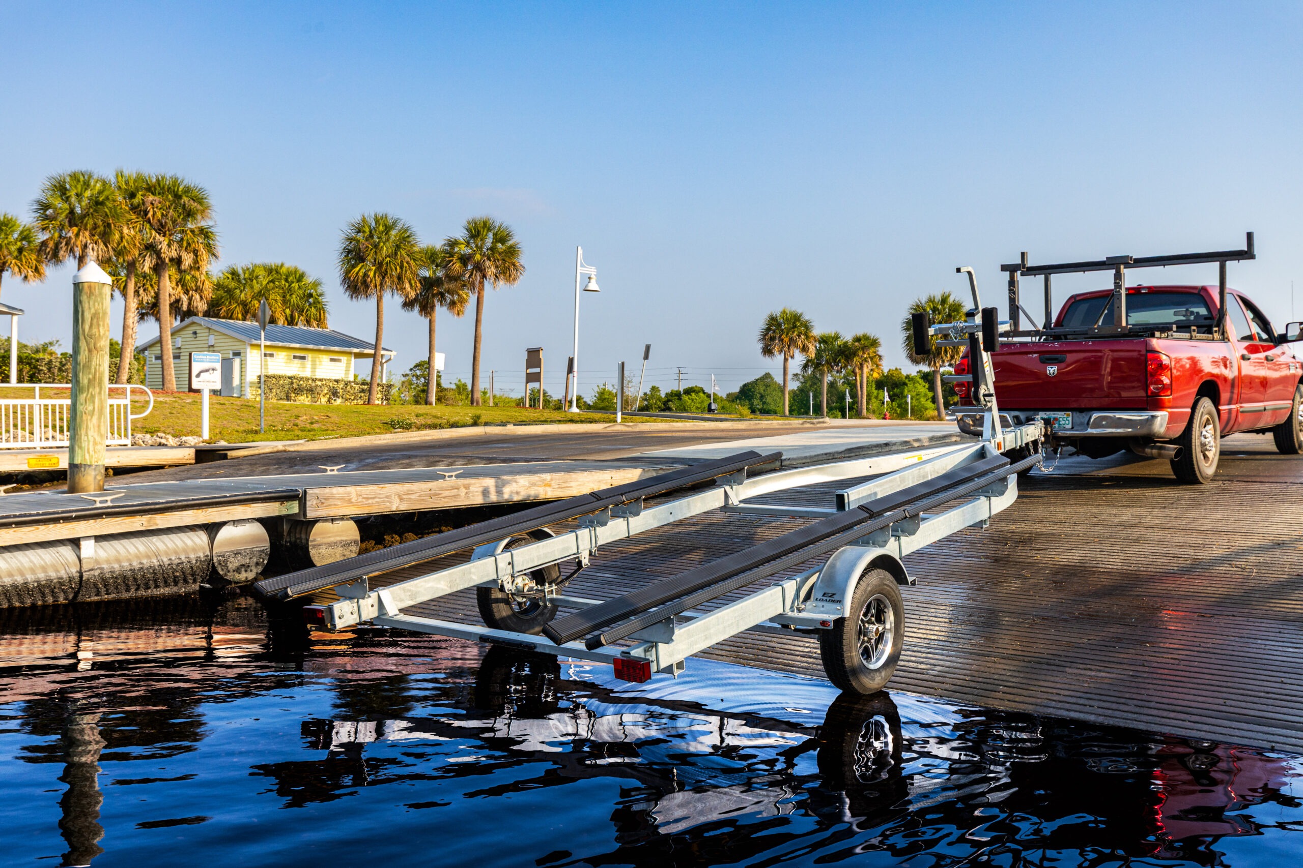 EZ Loader boat trailer backed into boat ramp with red pickup truck, galvanized frame and carpeted bunks visible over reflective water, ready for loading or launch