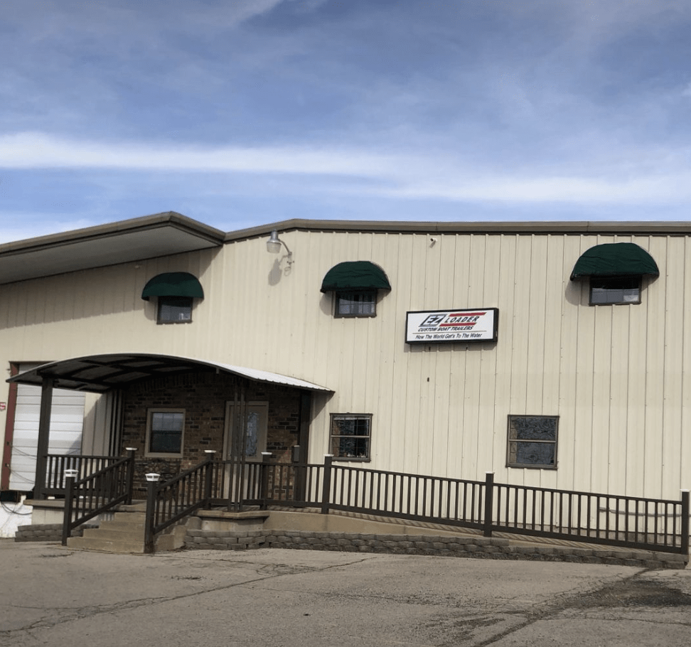 Screenshot 2025-09-19 at 5.00.48 PM Vintage photo of a small white and red boat on an early EZ Loader trailer in front of a workshop.